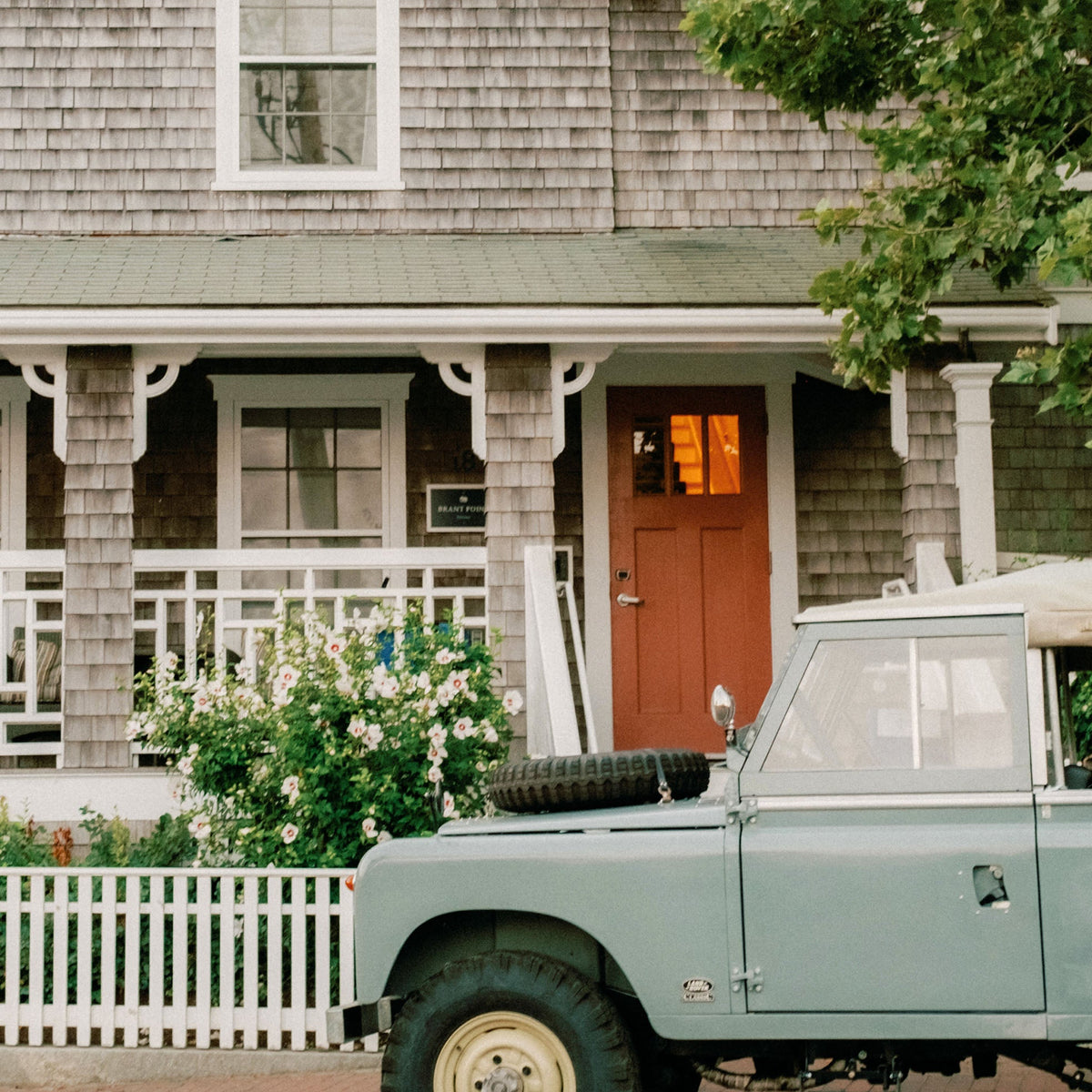 Close up of a vintage blue Land Rover parked in front of a house with a white picket fence and flowers.