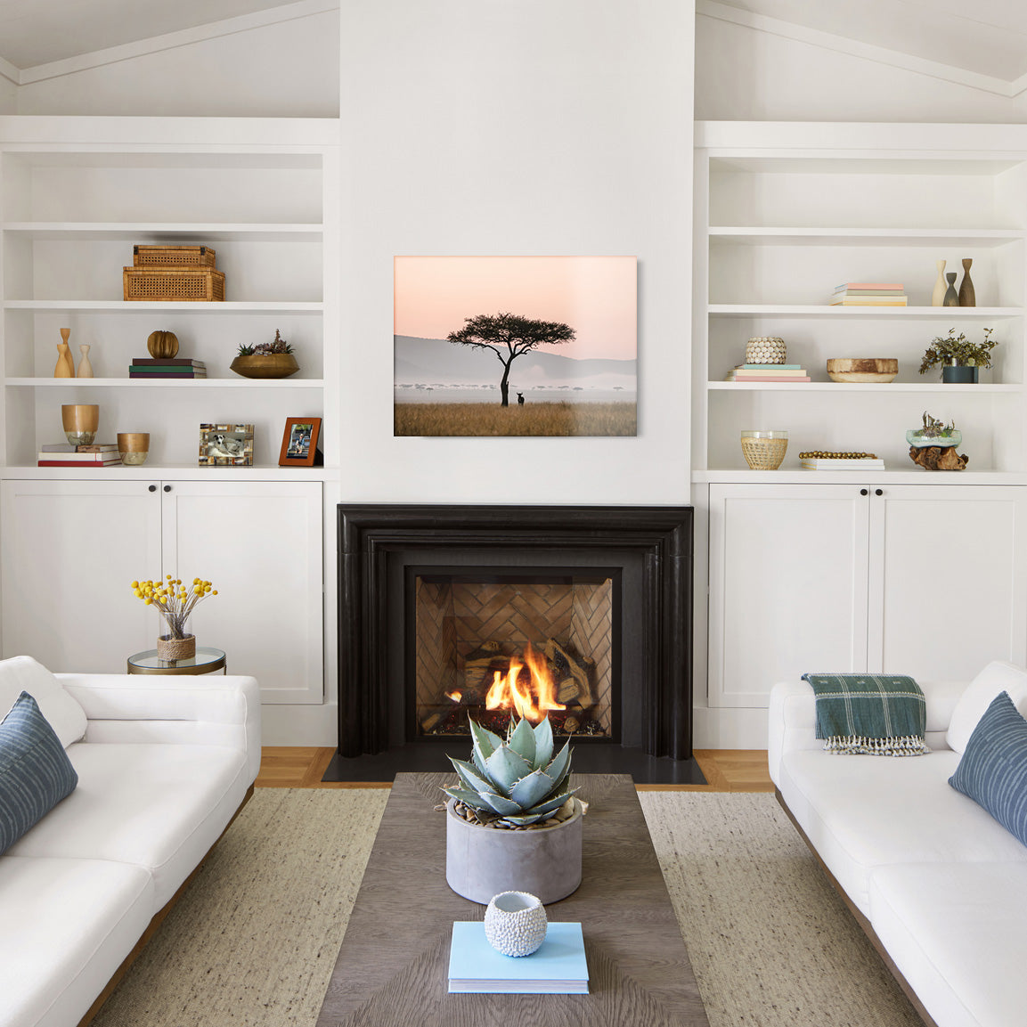 Modern living room with white walls, a fireplace, and a glass print of the silhouette of a tree and wildebeest against a pink sky.