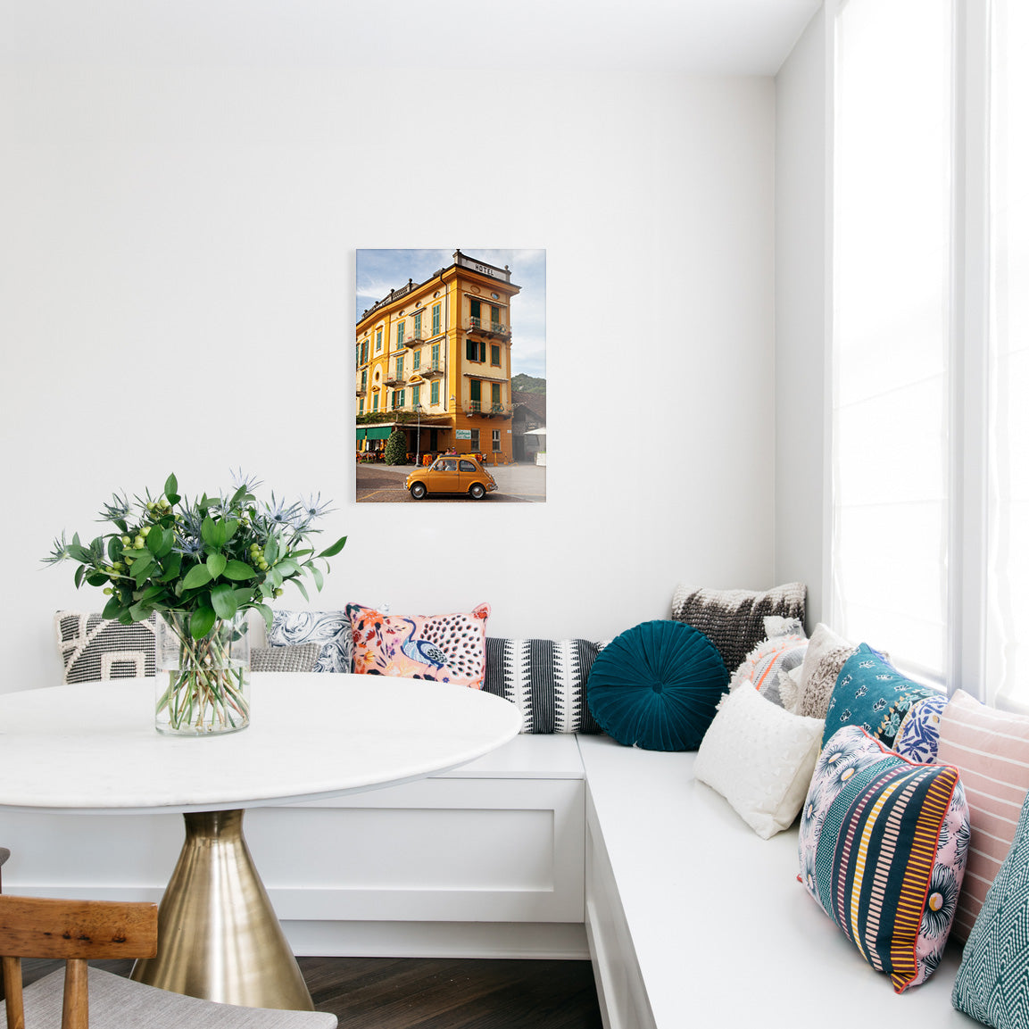 Kitchen nook with a white wall, colorful pillows on a sofa, a small table with flowers, and a glass print of a yellow vintage car in front of a yellow hotel.