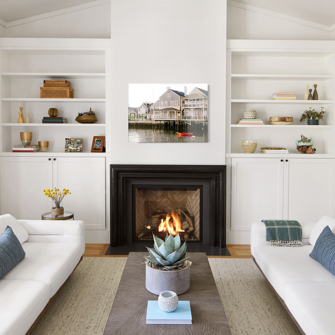 Living room with white walls, a fireplace, and a large glass print of a coastal house on the water with a red rowboat in the foreground.