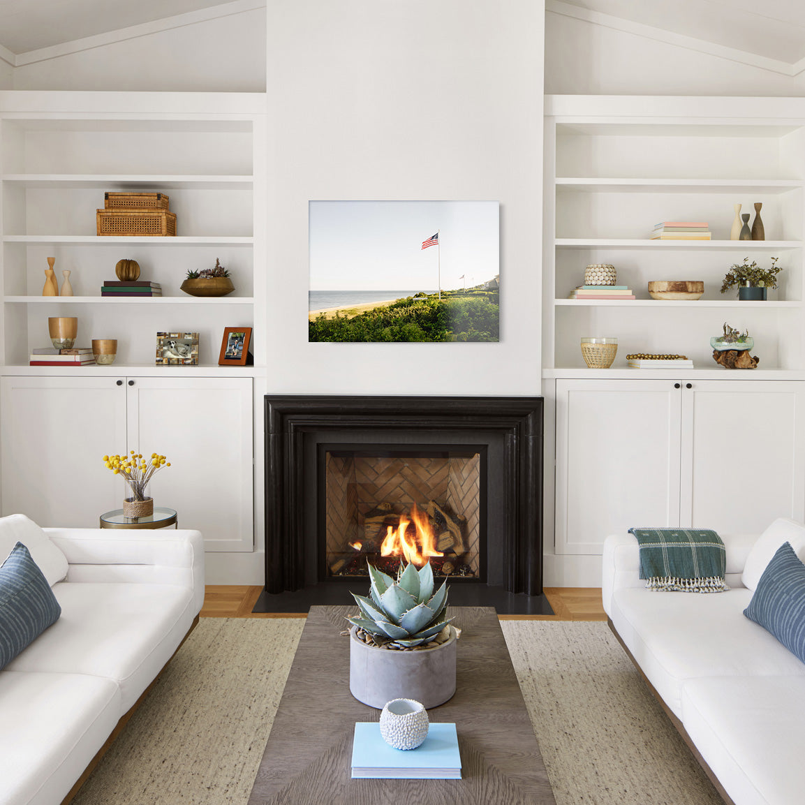Modern living room with white walls, a fireplace, and a glass print of a coastal view with a series of American flags on flagpoles along the water.