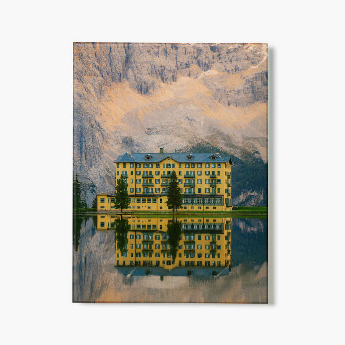 Glass Print of a yellow hotel, reflected in water in the foreground, with mountains in the background.