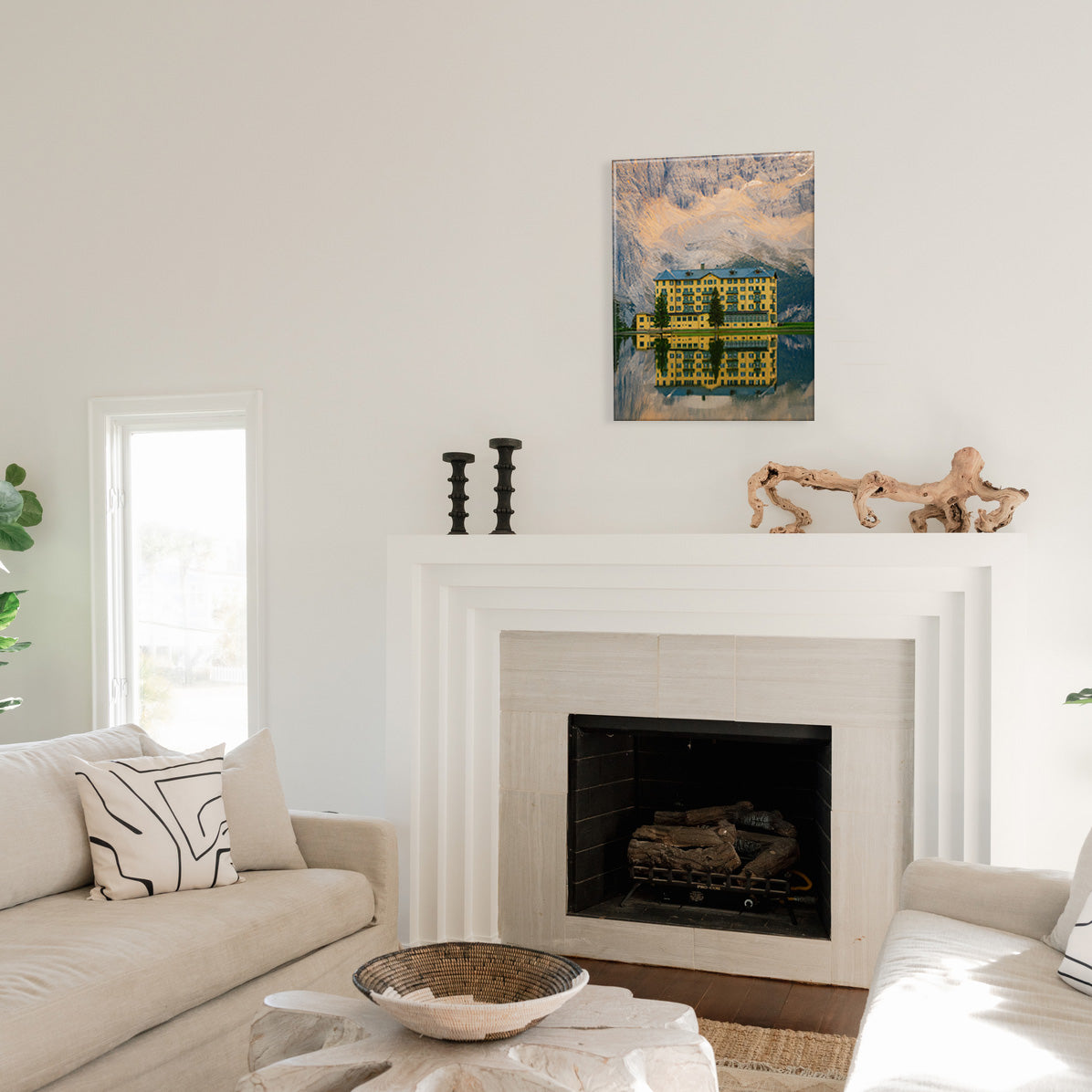 Living room with a fireplace and couch, and above the mantel hangs a glass print of a yellow hotel on the water's edge with a mountainous background.