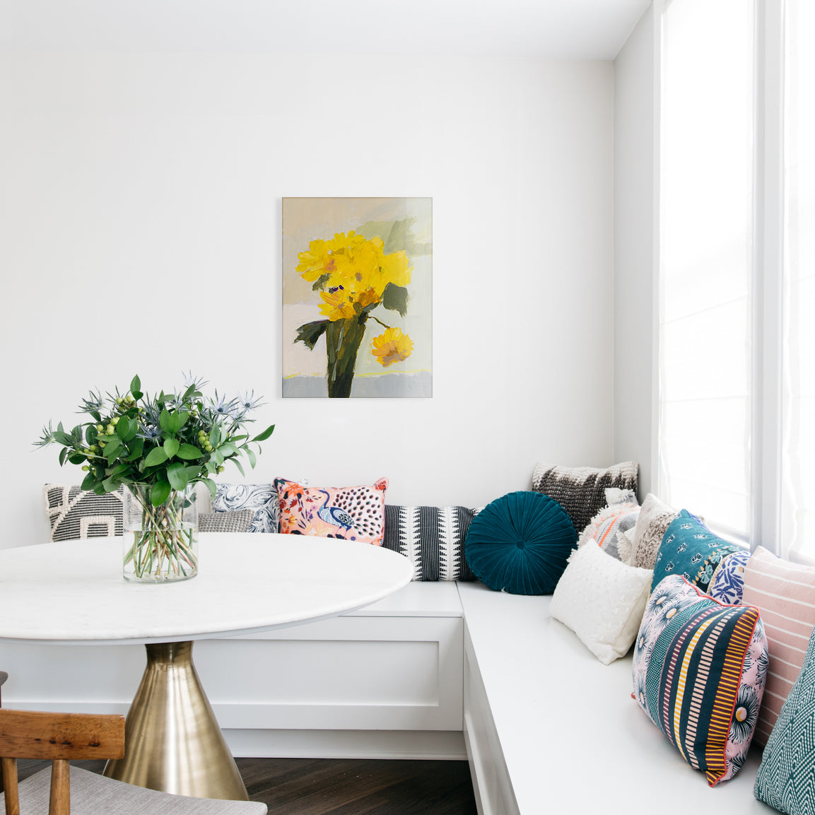 Kitchen with a white bench, colorful pillows, a small table with flowers, and a glass print of a painted yellow bouquet.